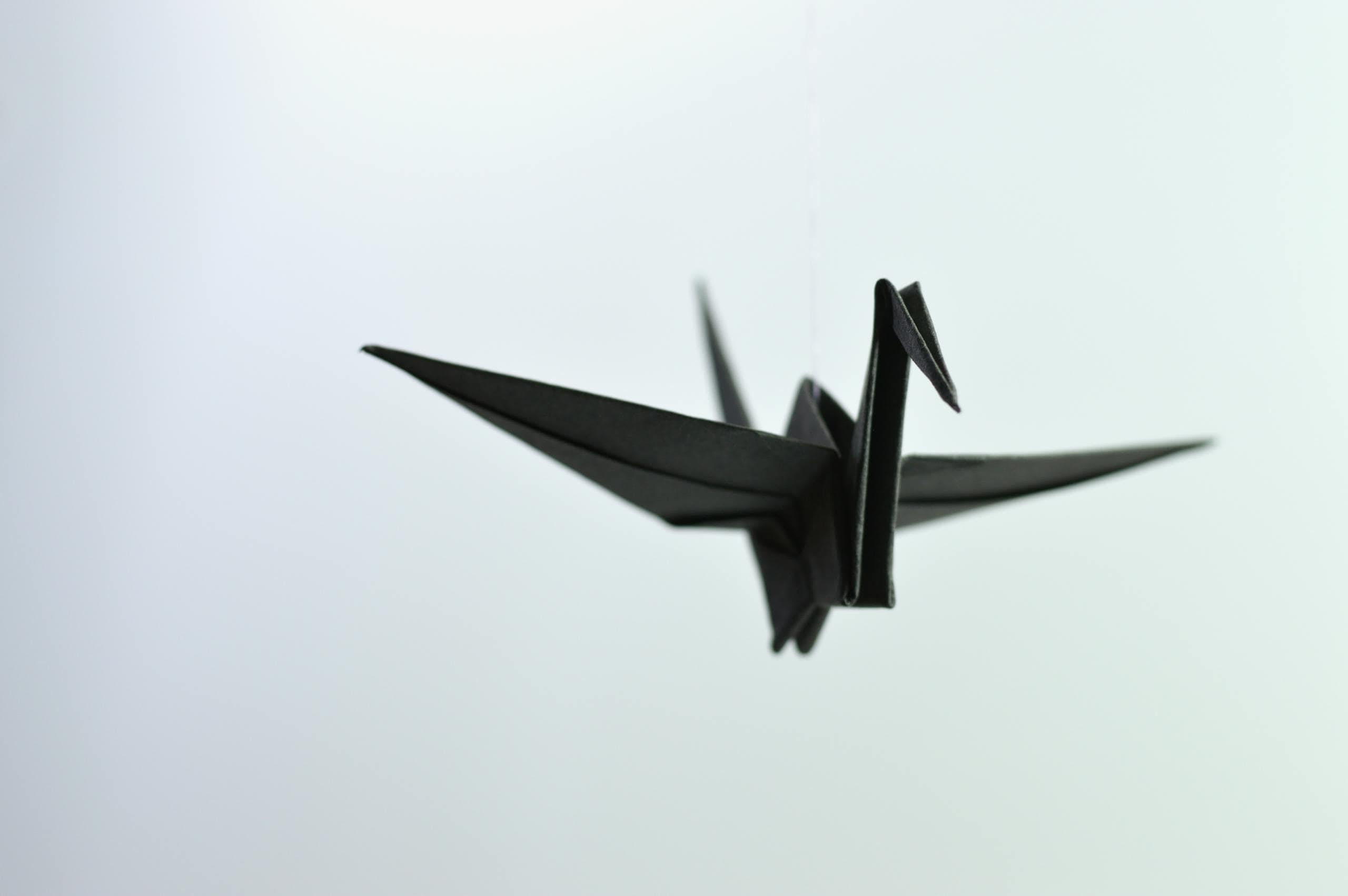 A minimalistic black origami paper crane hangs elegantly against a soft, blurred background, creating a serene atmosphere.