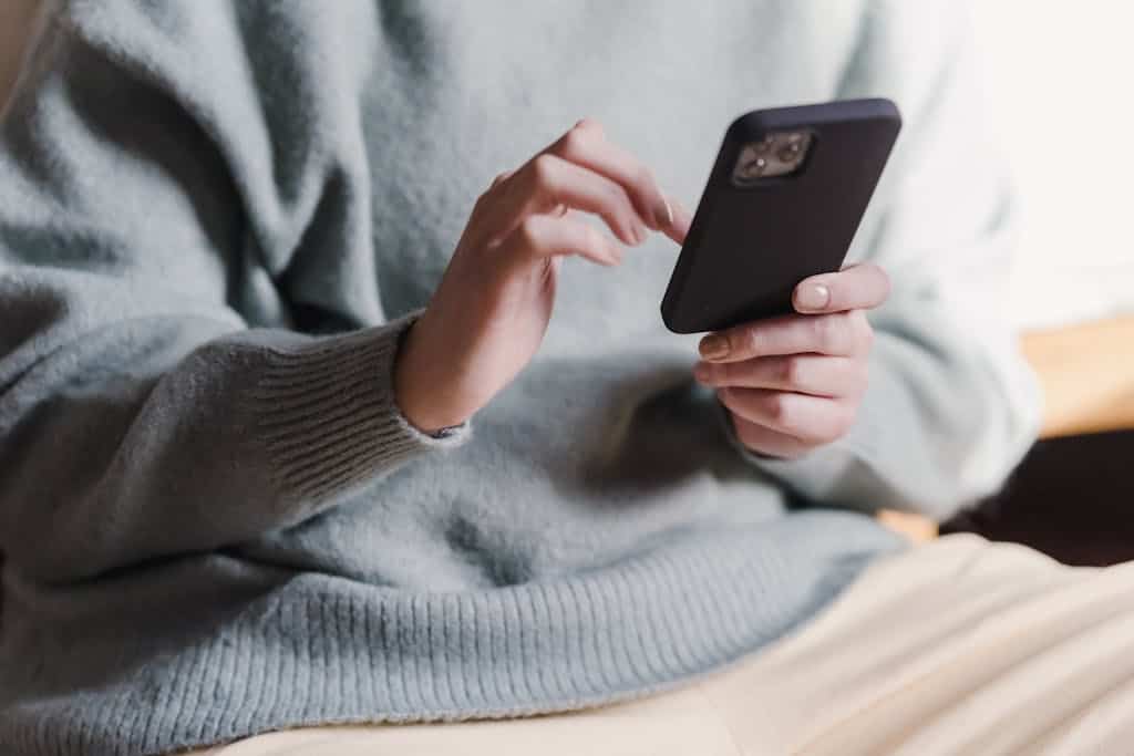 Crop faceless female in casual clothes using smartphone and touching screen while chilling at home