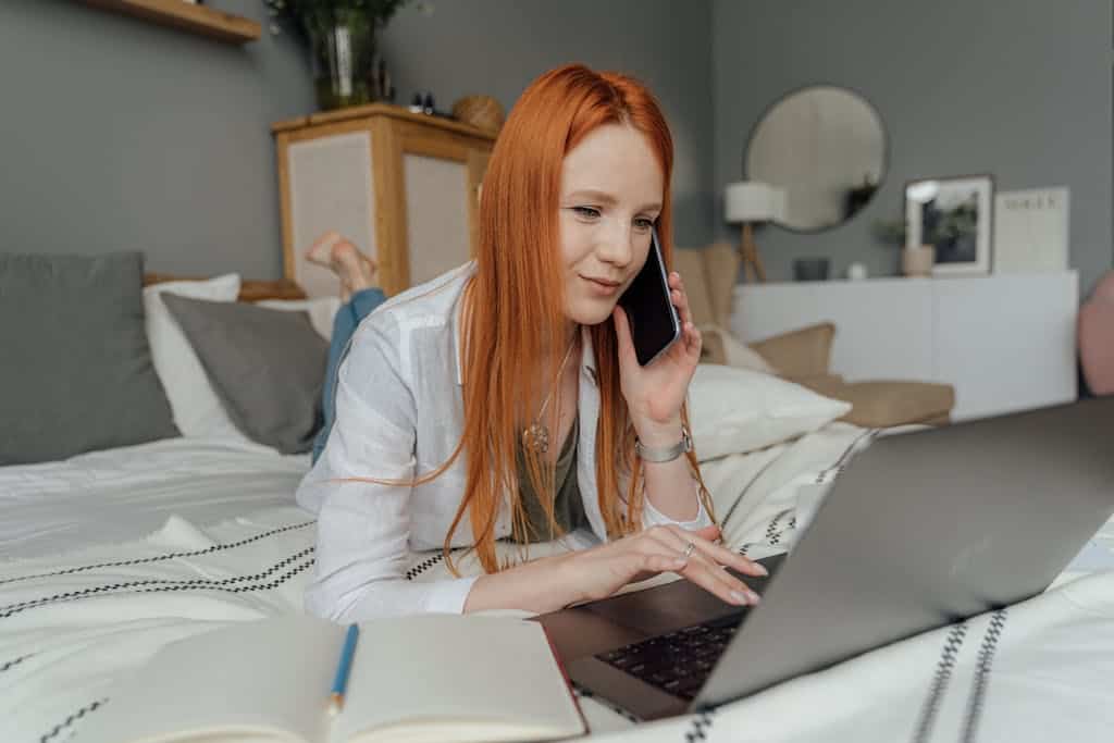 Rothaarige Frau bei der Webseitenerstellung: Sie arbeitet gleichzeitig am Laptop und am Smartphone in einem gemütlichen Schlafzimmer.